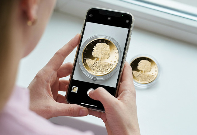 Frau scannt mit Smartphone die 100-Euro-Goldmünze Die Judenbuche